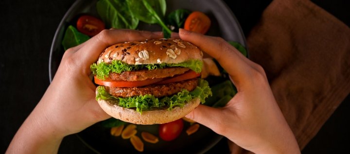 Zwei Hände halten einen vegetarischen Burger über einen Teller auf dem Salatblätter und Tomatenscheiben liegen.