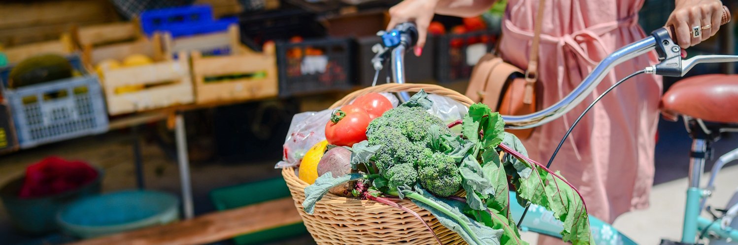 Eine Frau schiebt ein Fahrrad, an dessen Lenkrad ein Korb mit Gemüse hängt.