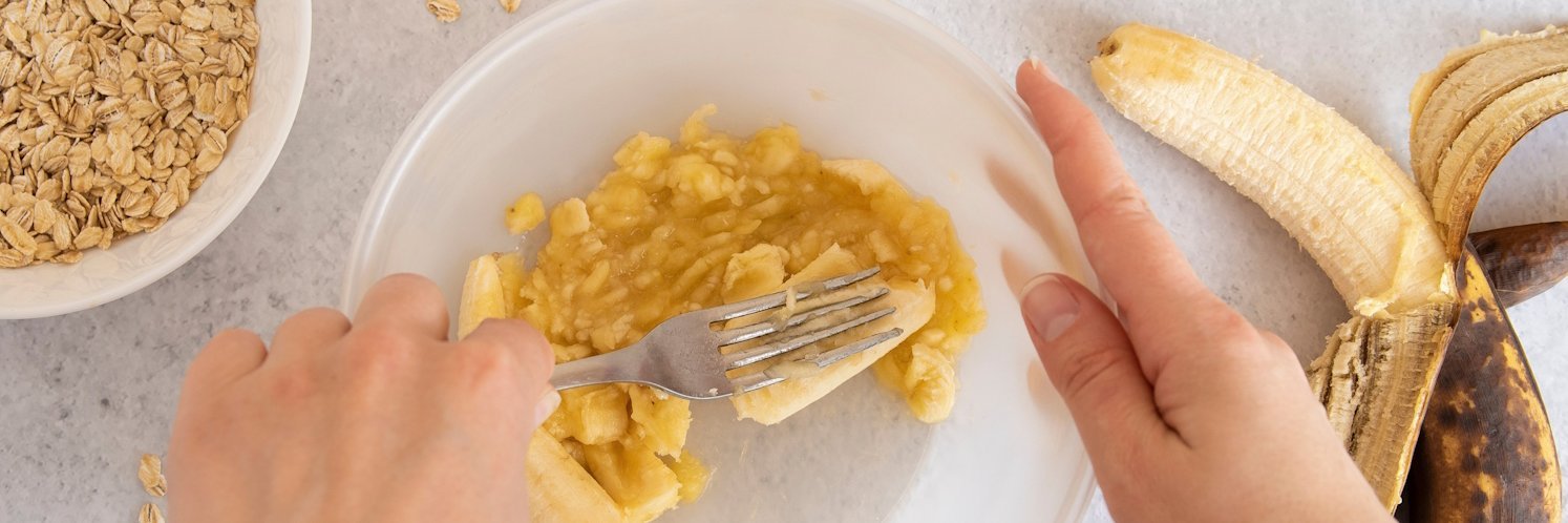 Hände einer Frau zerquetschen eine Banane mit einer Gabel in einem Teller.