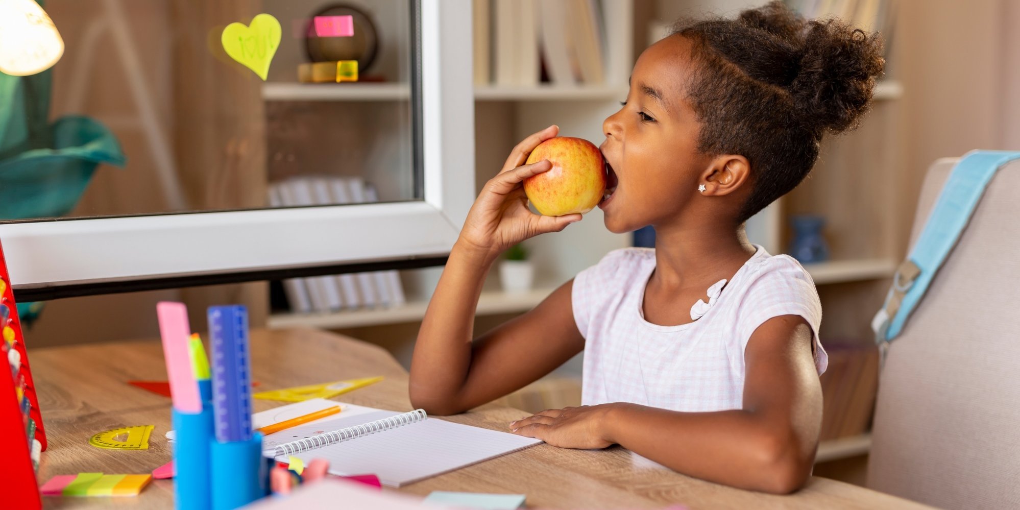 Eine Schülerin sitzt am Schreibtisch und beißt in einen Apfel.. Artikel "Was hat mein Apfel mit dem Klima zu tun?" öffnen.