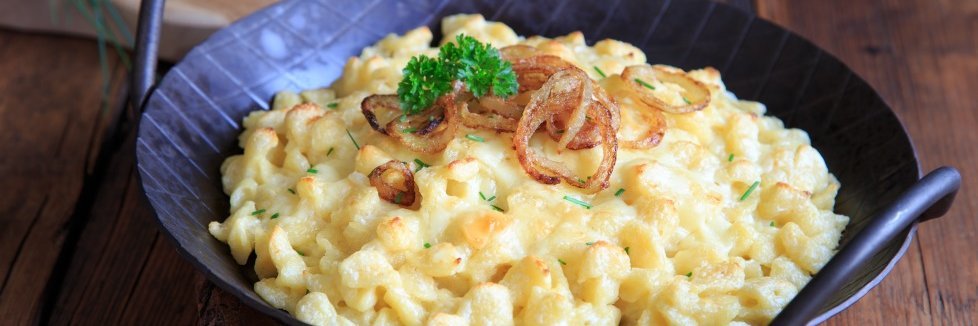 Auf einem dunklen Holztisch steht eine gusseiserne Pfanne mit Käsespätzle, dekoriert mit Petersilie und Röstzwiebeln.