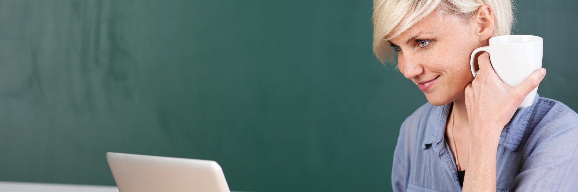 Eine Lehrerin sitzt am Tisch vor Tafel mit Laptop und Kaffeetasse.