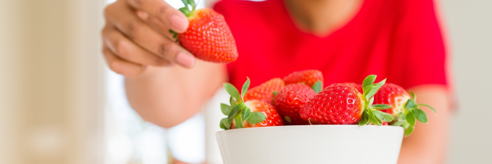Eine Frau mit rotem Pulli greift aus dem Hintergrund in eine Schale mit frischen Erdbeeren.