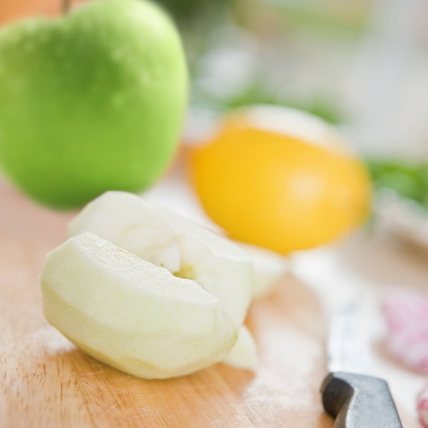 Auf einem Brett liegen ein halber geschälter Apfel, ein grüner Apfel, eine Zitrone und ein Küchenmesser.