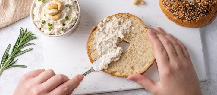 Auf einem weißen Brett bestreicht eine Person einen aufgeschnittenen Bagel mit veganem Cashew-Käse. Eine Schale mit dem Käse und einigen Cashewnüssen steht daneben.