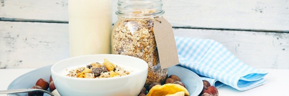 Apfel-Mandel-Müslimischung in einem Schraubglas. Daneben steht eine Flasche Milch und davor eine Schale mit Müsli und Milch. Ringsherum liegen Trockenfrüchte und Nüsse.