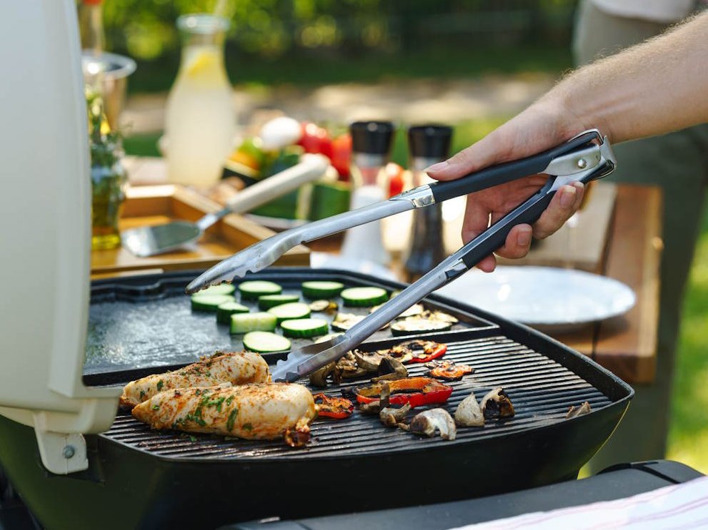Auf einem Grill liegt Fleisch und Gemüse. Eine Frau dreht mit einer Grillzange Fleisch um.