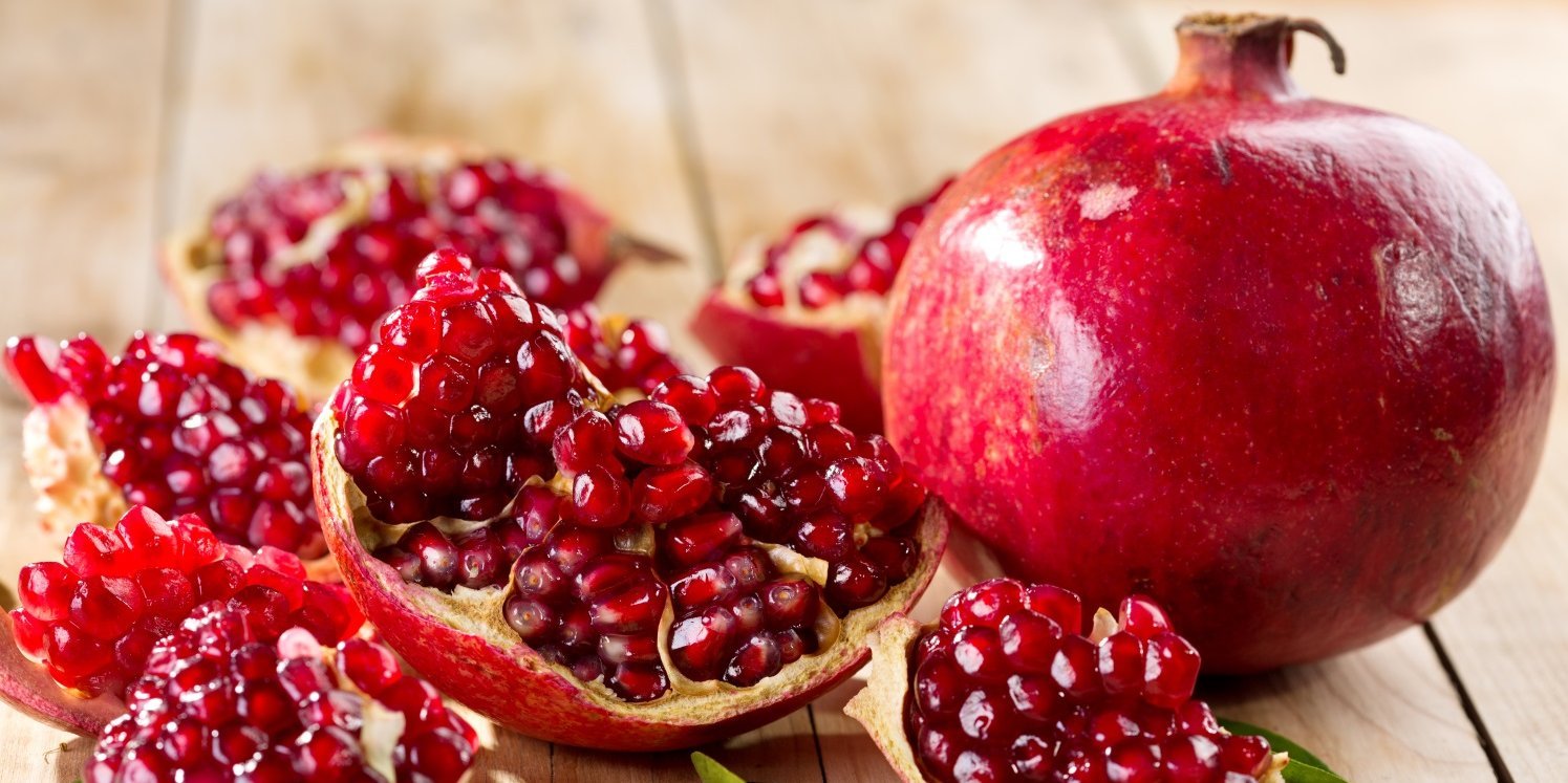 Auf einem hellen Holztisch liegt ein ganzer Granatapfel, daneben ein geöffneter in mehreren Stücken mit Samen darin.. Artikel "Kerniger Genuss: Der Granatapfel" öffnen.