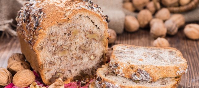 Auf einem dunklen Holztisch liegt ein aufgeschnittenes Kastenbrot mit Walnüssen im Teig, daneben liegen einzelne Scheiben und ganze Walnüsse.