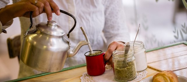 Eine Frauenhand gießt aus einem Wasserkessel Wasser in einen Becher aus dem ein Metalltrinkrohr ragt. Daneben stehen Behälter mit Mate-Blättern und Zucker auf einem Tisch.