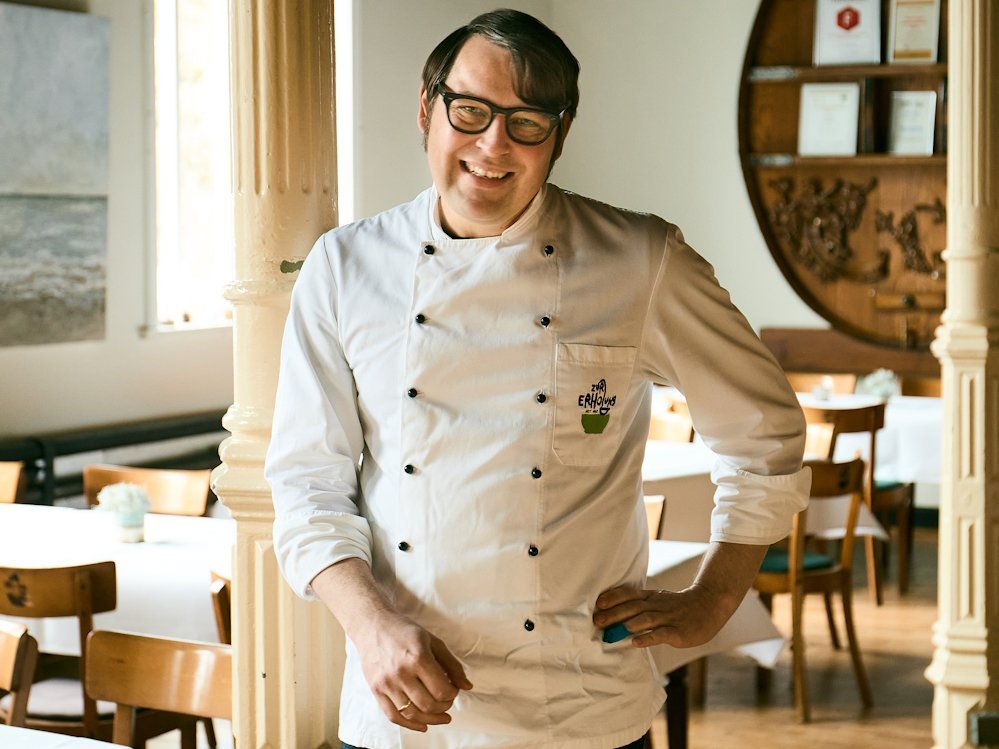 Ein Mann mit einer Kochjacke steht in einem Restaurant und lächelt in die Kamera.
