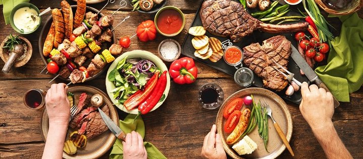 Auf einem Holztisch sind verschiedene gegrillte Speisen auf Platten und Tellern angerichtet. Neben Fleisch und Würstchen gibt es auch Gemüsespieße und Salat. Zwei Personen bedienen sich an der Auswahl.