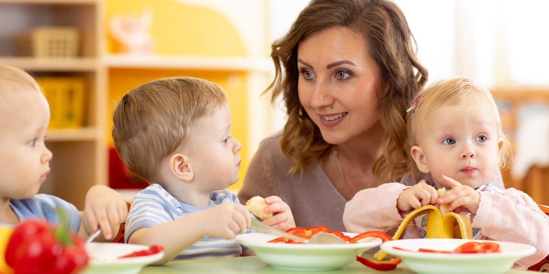 Eine Erzieherin sitzt zwischen zwei essenden Kleinkindern am Tisch.. Artikel "Partizipation beim Essen und Trinken" öffnen.