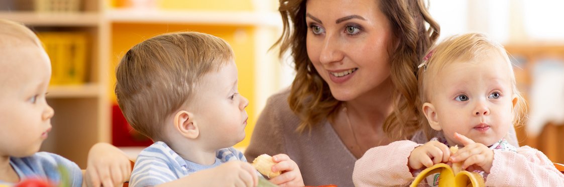 Eine Erzieherin sitzt zwischen zwei essenden Kleinkindern am Tisch.