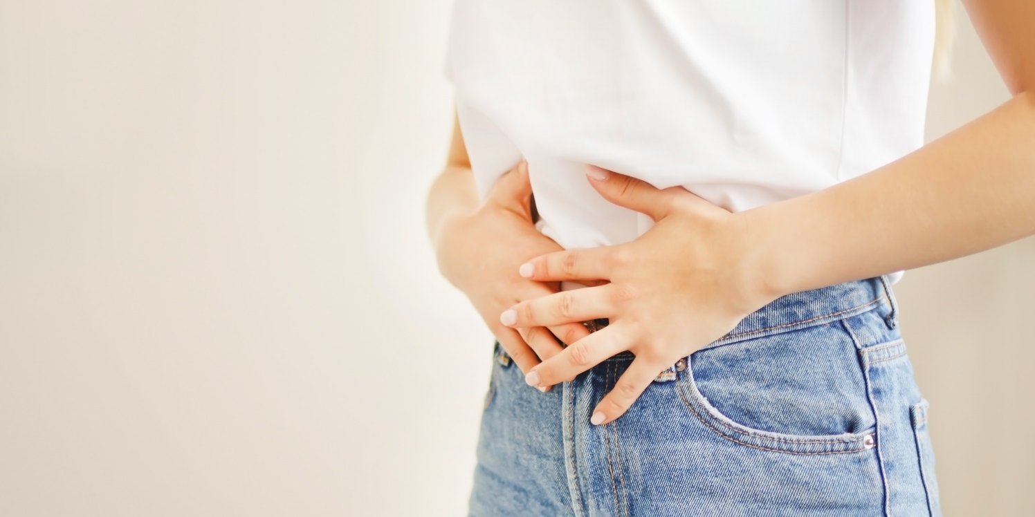 Eine Frau in Jeans und weißem T-Shirt drückt beide Hände auf ihren Bauch. . Artikel "Wie lange dauert eigentlich Verdauung?" öffnen.