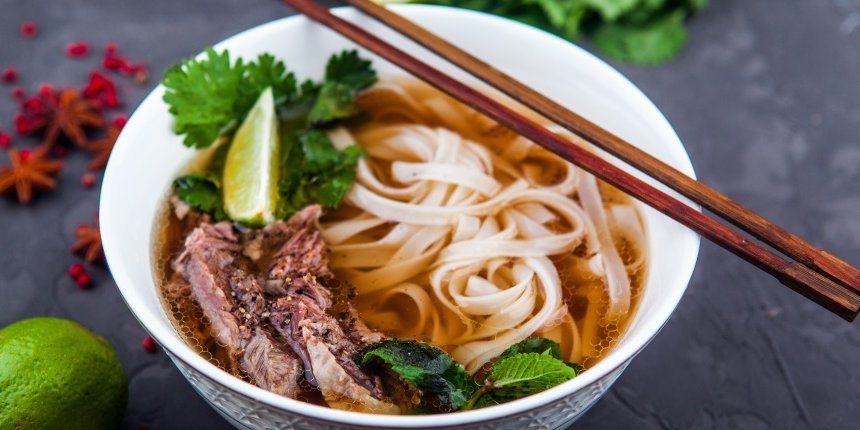 Auf einer dunklen Tischplatte steht eine weiße Suppenschüssel mit Pho-Suppe aus Reisbandnudeln, Fleisch und Kräutern. Auf dem Schüsselrand liegen Essstäbchen, daneben auf dem Tisch eine Limette, Sternanis und rote Pfefferkörner.. Artikel "Glasnudeln und Reisnudeln" öffnen.