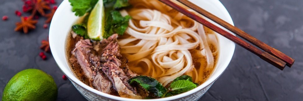 Auf einer dunklen Tischplatte steht eine weiße Suppenschüssel mit Pho-Suppe aus Reisbandnudeln, Fleisch und Kräutern. Auf dem Schüsselrand liegen Essstäbchen, daneben auf dem Tisch eine Limette, Sternanis und rote Pfefferkörner.