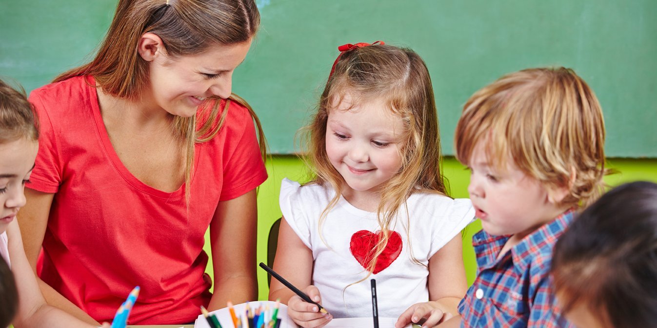 Eine Erzieherin sitzt mit Kita-Kindern am Tisch und malt. . Artikel "Ausmalbilder rund ums Essen" öffnen.