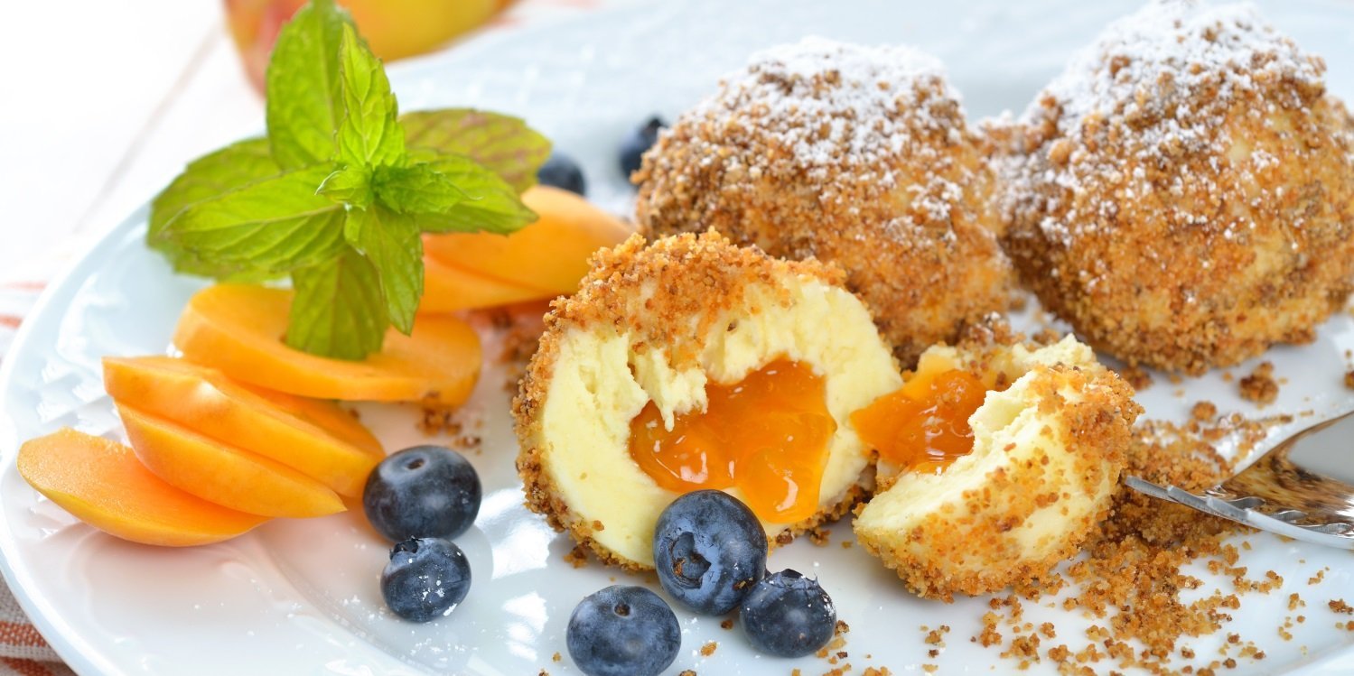 Auf einem weißen Teller liegen Marillenknödel mit Puderzucker. Einer ist halbiert und daneben liegen Aprikosenstücke und Blaubeeren.. Artikel "Die süß-saftige Aprikose" öffnen.