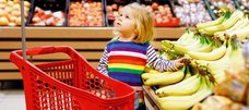 Ein etwa 3-jähriges Mädchen steht mit einem roten Kinder-Einkaufswagen in einer Obst- und Gemüseabteilung neben einer Auslage mit Bananen und schaut fragend nach oben.