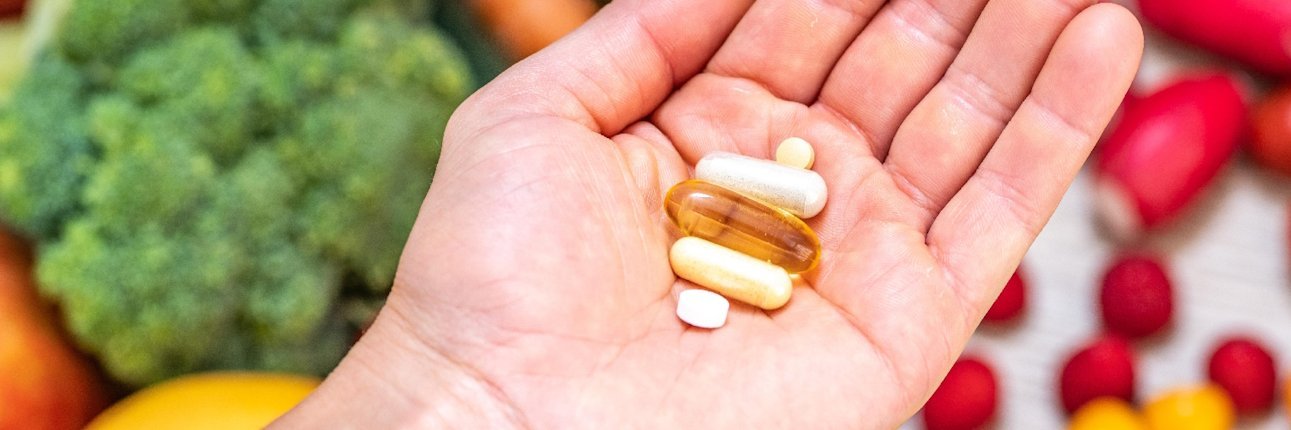 Auf einer Hand liegen mehrere Medikamente. Im Hintergrund sieht man diverses Gemüse und Obst.