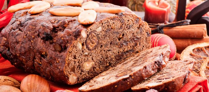 Auf einem weihnachtlich geschmückten Tisch mit Nüssen, Christbaumkugeln, Zimtstangen und Kerzen liegt ein dunkles Früchtebrot, das mit Mandeln verziert ist.  