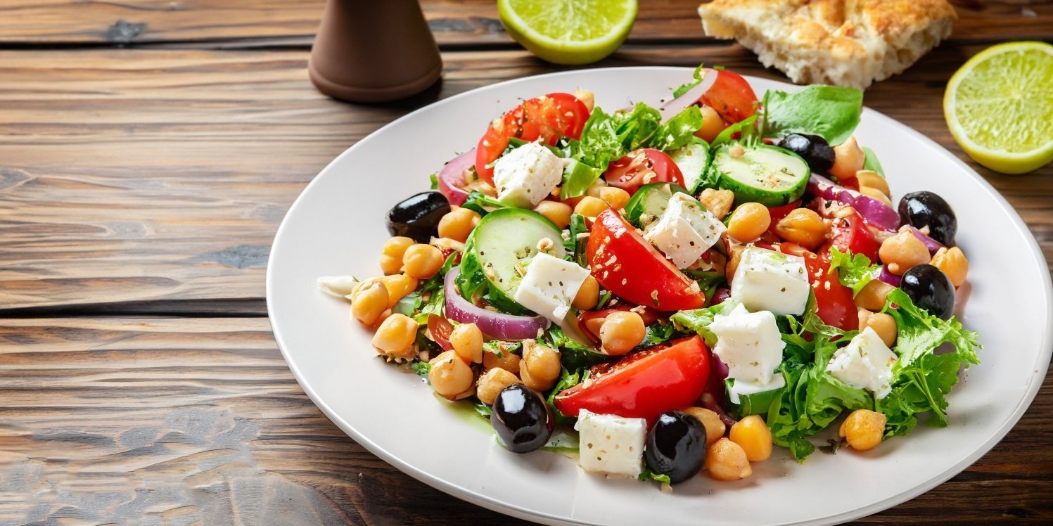 Auf einem dunklen Holztisch steht ein weißer Teller mit Blattsalat, Tomaten, Fetawürfeln, schwarzen Oliven und Kichererbsen.. Artikel "Kichererbsen in der Küche" öffnen.