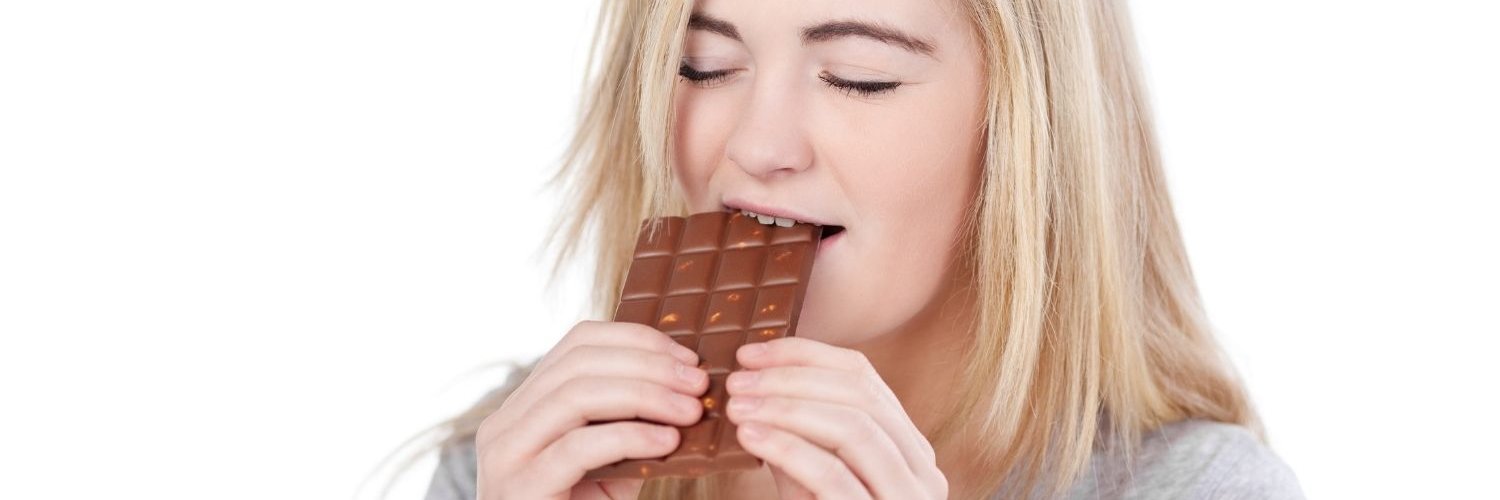 Eine junge blonde Frau beißt mit geschlossenen Augen in eine ganze Tafel Schokolade.