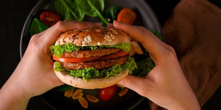 Zwei Hände halten einen vegetarischen Burger über einen Teller auf dem Salatblätter und Tomatenscheiben liegen.. Artikel "Mehr Fleischalternativen auf dem Teller" öffnen.