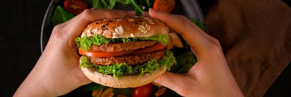 Zwei Hände halten einen vegetarischen Burger über einen Teller auf dem Salatblätter und Tomatenscheiben liegen.