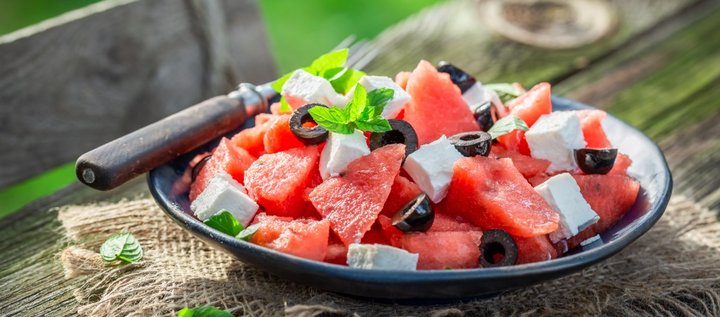 Auf einem Holztisch im Freien steht ein dunkler Teller mit gewürfelter Wassermelone, Fetastücken, schwarzen Oliven und Minzblättern. 