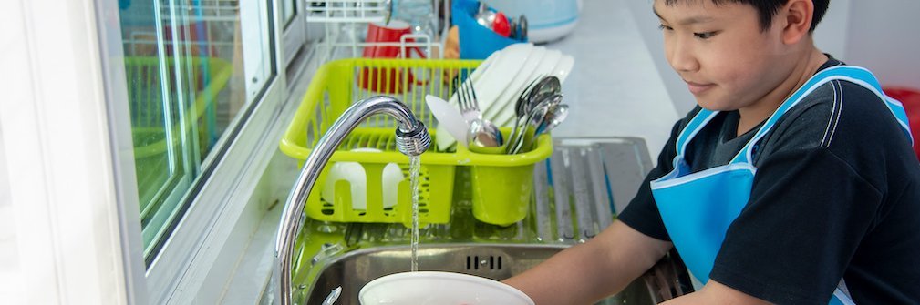 Ein Junge mit einer blauen Schürze spült Teller am Waschbecken ab.