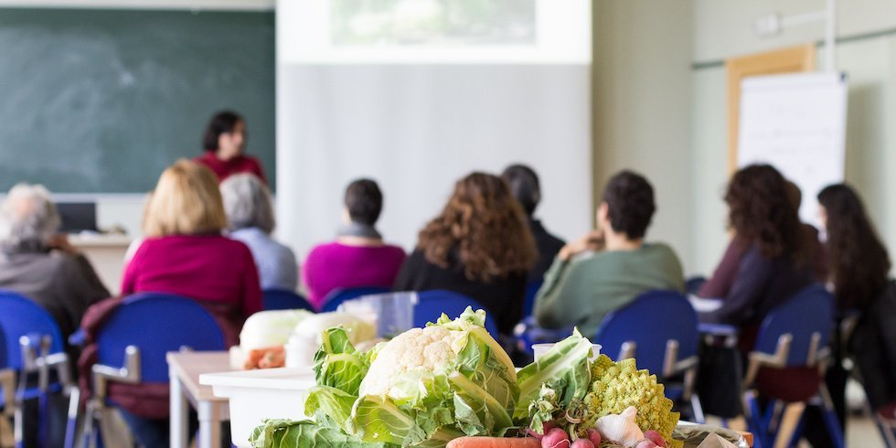 Erwachsene sitzen in einem Klassenzimmer, auf einem Tisch liegt Gemüse.. Artikel "Erwachsene" öffnen.