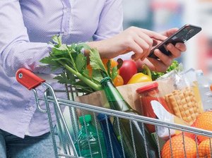 Eine Frau mit einem Handy in der Hand lehnt auf einem Einkaufswagen mit Lebensmitteln.