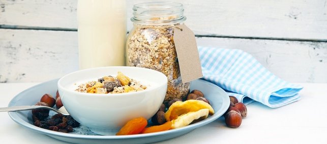 Apfel-Mandel-Müslimischung in einem Schraubglas. Daneben steht eine Flasche Milch und davor eine Schale mit Müsli und Milch. Ringsherum liegen Trockenfrüchte und Nüsse.