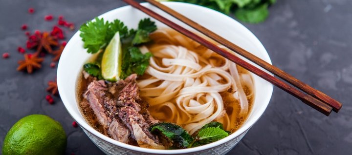 Auf einer dunklen Tischplatte steht eine weiße Suppenschüssel mit Pho-Suppe aus Reisbandnudeln, Fleisch und Kräutern. Auf dem Schüsselrand liegen Essstäbchen, daneben auf dem Tisch eine Limette, Sternanis und rote Pfefferkörner.