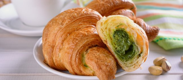 Auf einem Teller liegt ein aufgeschnittenes Croissant mit Pistaziencreme gefüllt. Daneben liegen einige Pistazien und eine gestreifte Serviette neben einer Kaffeetasse.