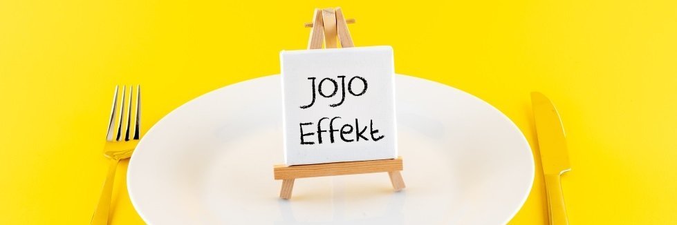 Auf einem gelben Untergrund steht ein weißer Teller, daneben liegt Besteck. Auf dem Teller steht eine Mini-Staffelei mit einem Schild mit der Aufschrift „Jojo-Effekt“.