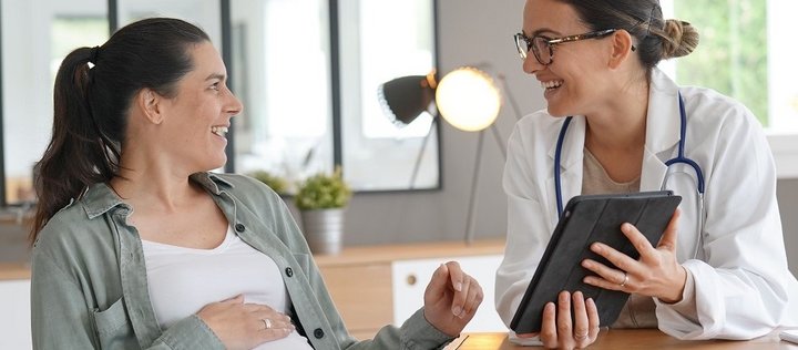 Eine schwangere Frau sitzt mit einer Ärztin an einem Tisch. Die Ärztin hält ein Tablet in der Hand. Beide schauen sich an und wirken fröhlich. 