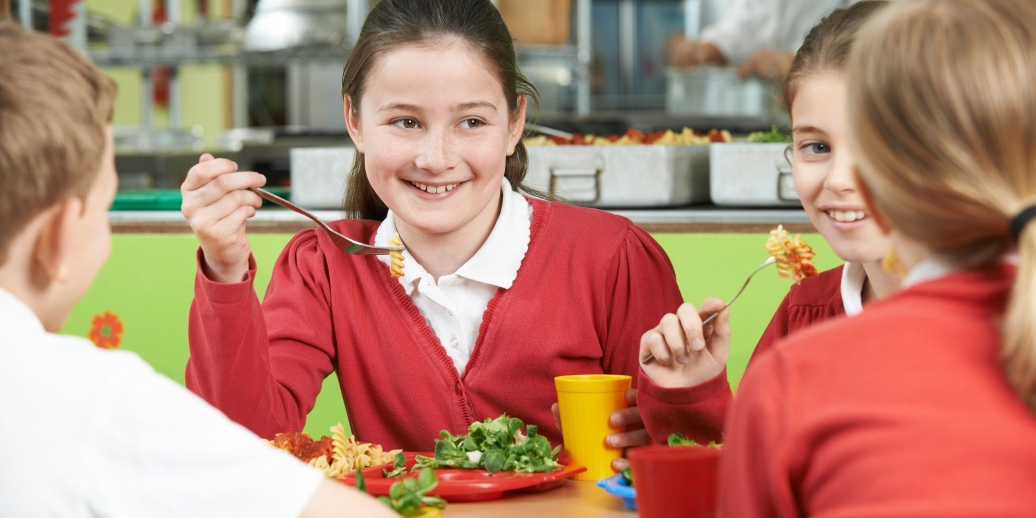 In einer Kantine sitzen vier Kinder miteinander am Tisch und essen ein Nudelgericht und Salat. . Artikel "Mehr Kinder für kostenfreies Schulmittagessen gewinnen" öffnen.