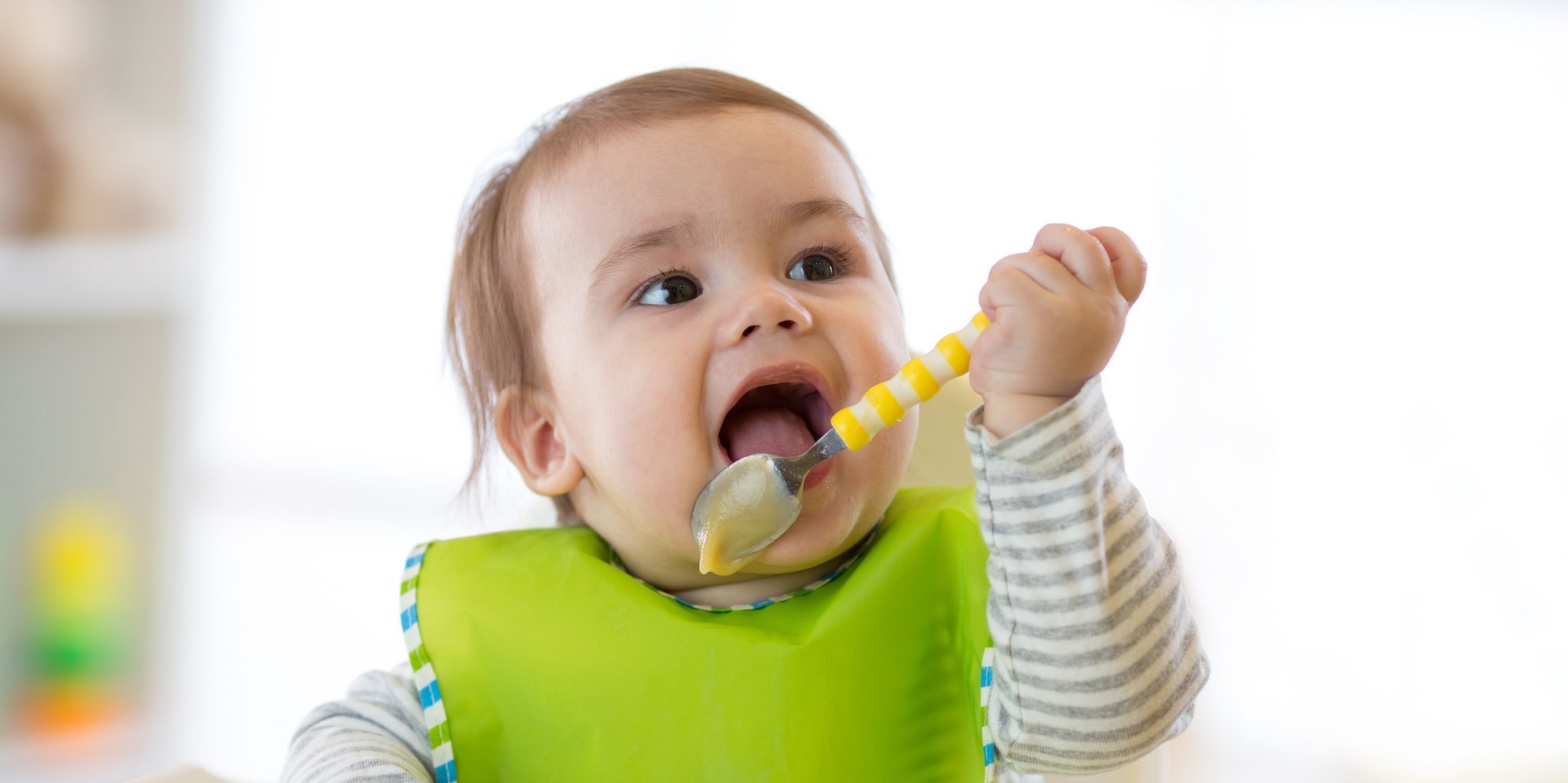 Ein Kleinkind mit Lätzchen leckt Brei vom Löffel ab.. Artikel "Ernährungsbildung in der Kindertagespflege" öffnen.