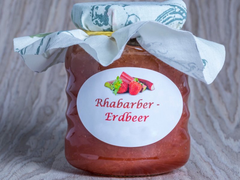 Ein Glas selbstgemachte Erdbeer-Rhabarber-Marmelade steht auf hellgrauem Hintergrund.