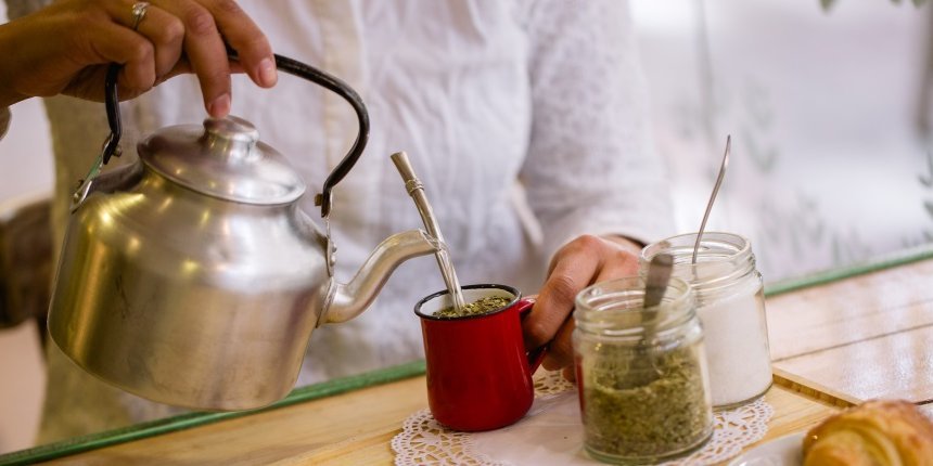 Eine Frauenhand gießt aus einem Wasserkessel Wasser in einen Becher aus dem ein Metalltrinkrohr ragt. Daneben stehen Behälter mit Mate-Blättern und Zucker auf einem Tisch.. Artikel "Mate-Tee richtig zubereiten" öffnen.