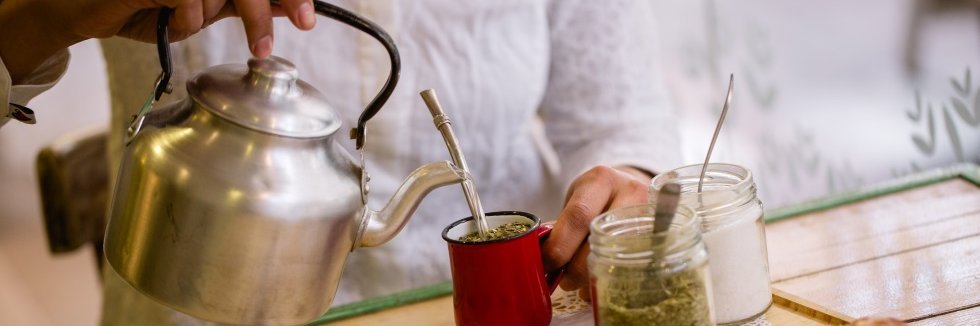 Eine Frauenhand gießt aus einem Wasserkessel Wasser in einen Becher aus dem ein Metalltrinkrohr ragt. Daneben stehen Behälter mit Mate-Blättern und Zucker auf einem Tisch.