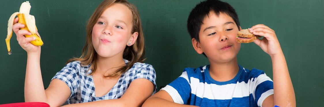 Zwei Schulkinder sitzen vor einer Tafel und essen ein Brot und eine Banane.