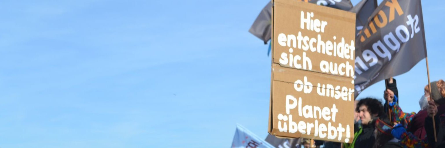 Schild auf einer Demonstration am Rand einer Kohlegrube in Lützerath mit der Aufschrift "Hier entscheidet sich auch, ob unser Planet übelebt".  