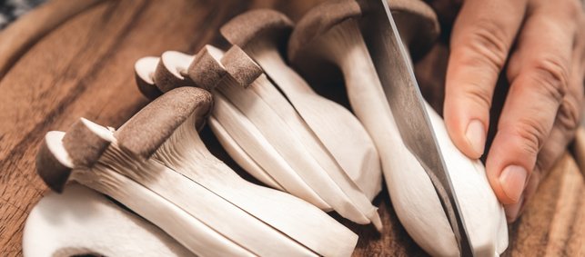 Auf einem dunklen Schneidebrett liegen mehrere Kräuterseitlinge, längs in Streifen geschnitten. Eine Hand mit Messer schneidet einen weiteren Pilz längs durch.