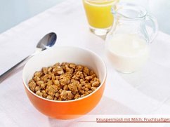 Schale mit Knuspermüsli, Löffel, Glas Saft und Krug mit Milch.