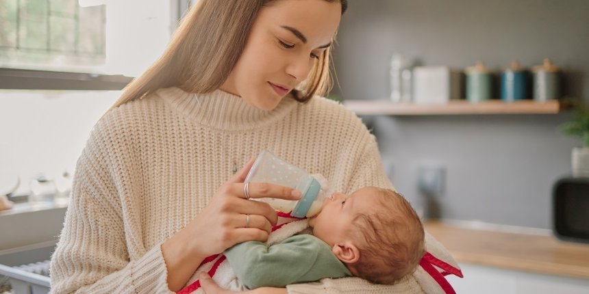 Eine Frau steht in einer Küche mit einem Baby auf dem Arm und füttert es mit einem Fläschchen.. Artikel "Wie Flaschennahrung richtig gefüttert wird" öffnen.