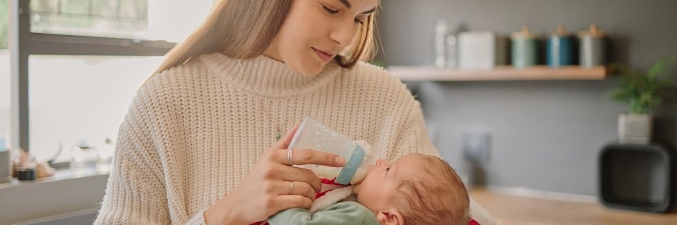 Eine Frau steht in einer Küche mit einem Baby auf dem Arm und füttert es mit einem Fläschchen.